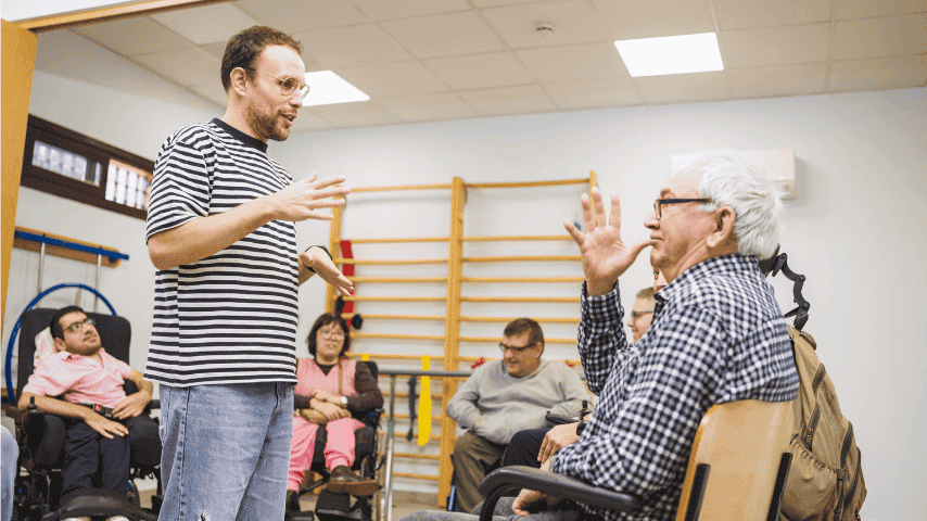 A Deaf man communicating in sign language with another man during an inclusive group discussion, with disabled participants seated in the background.