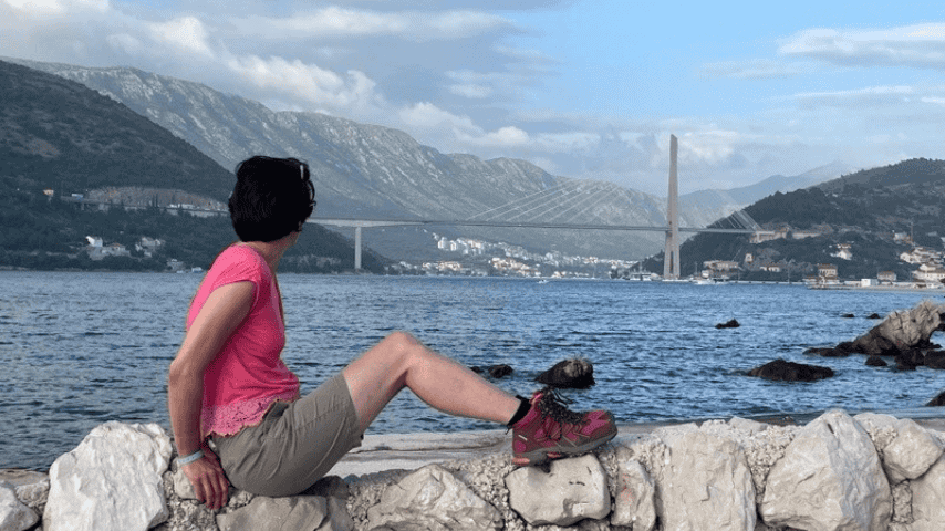 A woman sitting on a stone wall looking across the water towards a bridge and mountains on the Adriatic coast.