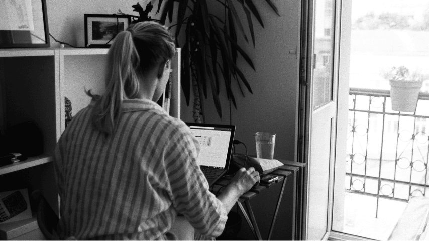 A woman sitting at a laptop beside an open balcony door, representing the often unseen administrative work involved in navigating healthcare, education and support systems.