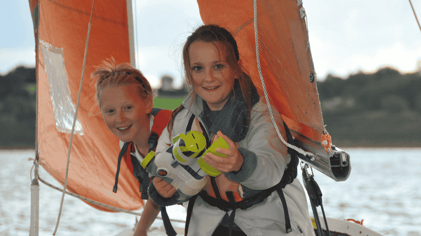 Two young people smiling aboard a small sailing dinghy, wearing buoyancy aids with an orange sail behind them.
